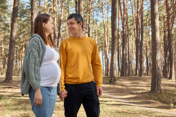 Obraz premium Cheerful smiling man and woman standing in park holding hands looking at each other husband and his pregnant wife walking in spring forest admiring nature and breathing fresh air