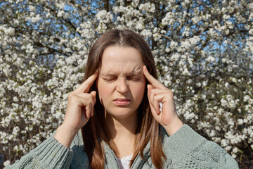 Sad upset young woman having allergy symptoms from blooming tree pollen in spring suffering...