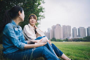 Two Asian women chat and enjoy outdoor time in a city park. Amidst the urban setting, they engage in conversation, savoring the moments spent outdoors in the park.