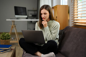 Fototapeta na wymiar Attractive young woman in casual clothes suing laptop sitting on couch at home