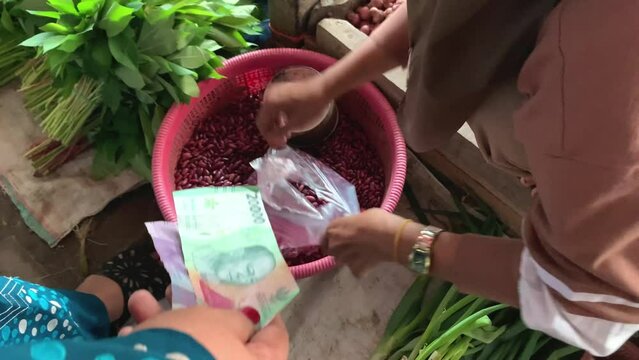 A seller put red beans in a plastic bag and a buyer pays in Indonesian currency. Transaction on traditional market.
