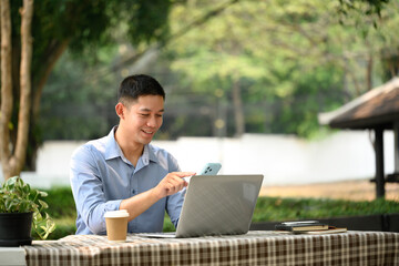Smiling adult asian man using mobile phone and working with laptop at outdoor cafe