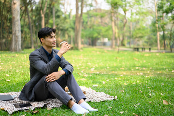 Handsome young businessman sitting on meadow and drinking coffee from paper cup