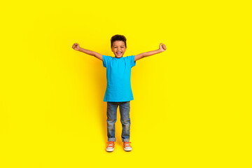 Full body photo of cheerful overjoyed small child dressed blue t-shirt jeans flying clenching fists isolated on vibrant yellow background