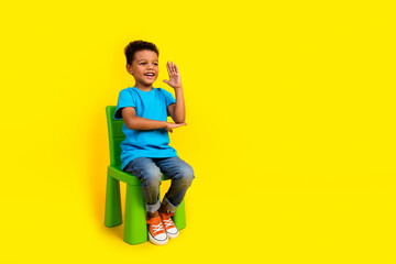 Full body photo of cute small child dressed blue t-shirt sit on chair look empty space raising palm...