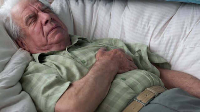 Elderly gray haired caucasian male patient lay down on bed in house feeling unwell and having difficulty breathing. Old man has strong sharp pain in chest puts hand on heart. Suffers from heart attack