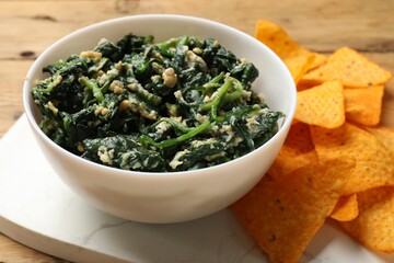 Tasty spinach dip with eggs in bowl and nachos chips on wooden table, closeup