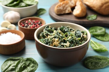 Tasty spinach dip with egg in bowl and spices on light blue wooden table, closeup