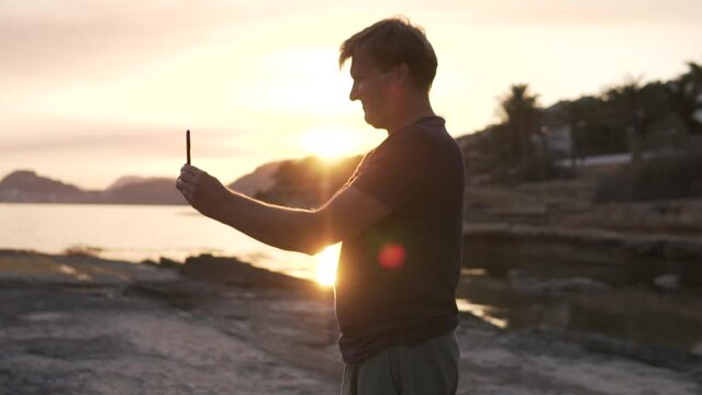 A man takes a photo of the sea at sunset on a mobile phone.