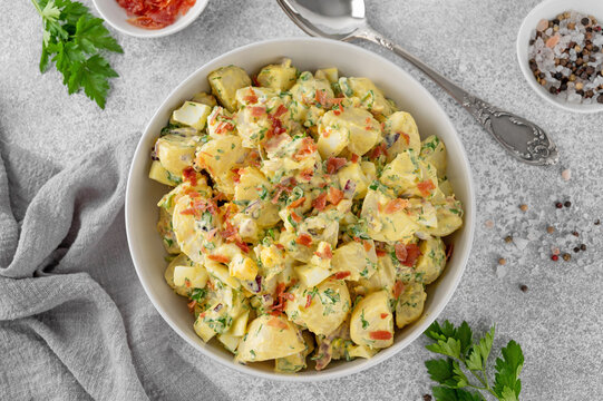 Salad of potatoes, eggs, bacon and fresh herbs with mayonnaise and yogurt dressing in a bowl on a gray concrete background.