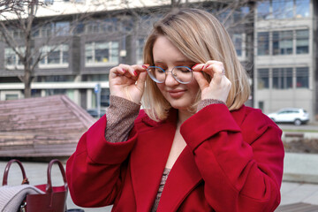Young woman, blonde in red coat, sitting on bench in city, uses glasses to see better. Vision...