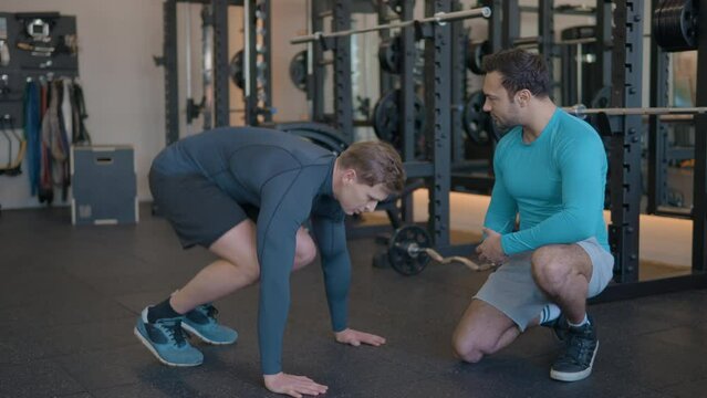 Personal coach supervises man working out in gym doing plank jumps in slow motion.