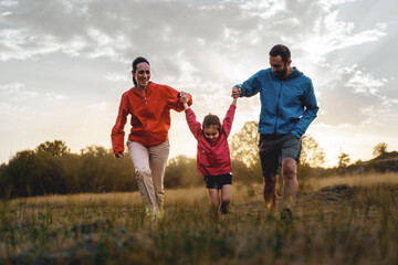 Family enjoys a playful run in the field at sunset, with a child swinging between parents,...
