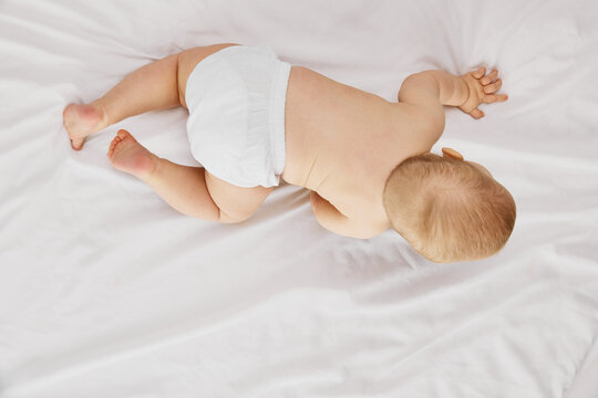 Aerial view photo of little charming baby lies on belly, on comfy, soft bed covered with white fabric. Cute explorer. Concept of childhood, motherhood, life, birth. Copy space for ad