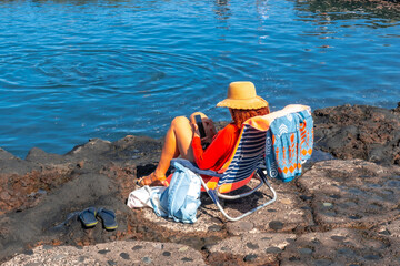 A woman is sitting in a chair by the water, wearing a straw hat