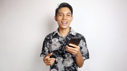 Happy young handsome Asian man wearing batik shirt posing holding smartphone
