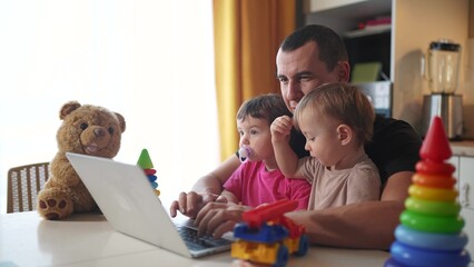 father working from home a remotely with two fun baby in his arms. pandemic remote work business concept. father tries to work at home in kitchen, baby children interfere sitting on their hands