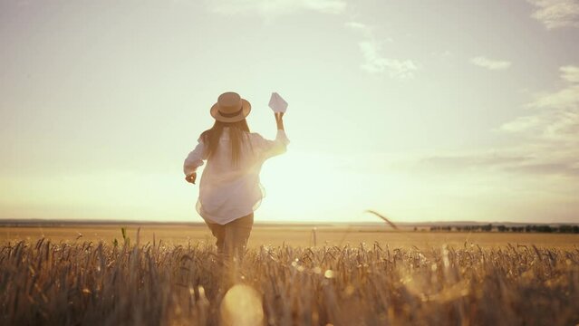 Carefree woman running on wheat field throwing paper plane in sky keeping straw hat on head at sunset, back view. Freedom, farm lifestyle, playful in countryside village, outdoor activities concept.