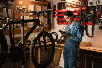 Young cute woman repair bicycle with mobile phone search spare parts on the internet in garage workshop. diy concept