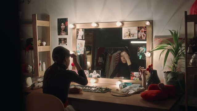 Female mime artist sitting in dressing room at vanity desk, looking in mirror and applying white face paint while doing stage makeup
