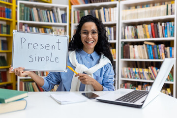 Cheerful female teacher holding a board with 'Present Simple' written on it, demonstrating English...