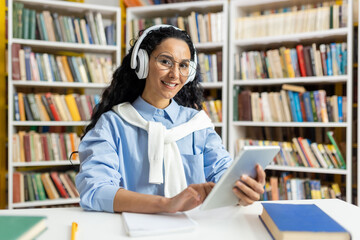A cheerful woman wearing headphones is comfortably using a digital tablet in a well-stocked library, surrounded by numerous books.