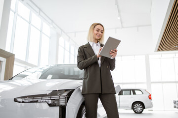 Professional smiling female car dealer posing at auto showroom holding tablet pc. Happy saleswoman...