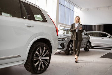 Professional Caucasian female dealer with digital tablet smiling and looking at camera friendly. Young saleswoman standing near modern automobile in bright car showroom.