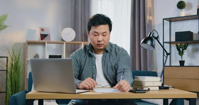 Asian man working on laptop sitting at his workplace at home writing out needed datas on paper for his business project. Learning and education concept.