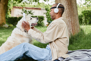A disabled African American man with myasthenia gravis syndrome sits in the grass with a Labrador dog wearing headphones, enjoying a peaceful moment together.