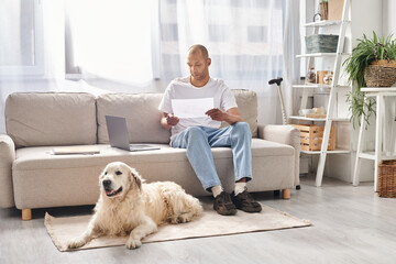 A disabled African American man with myasthenia gravis sits on a couch next to his Labrador dog at...