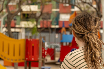 Fototapeta premium Child exploitation: Blond-haired boy with dreadlocks watches playground