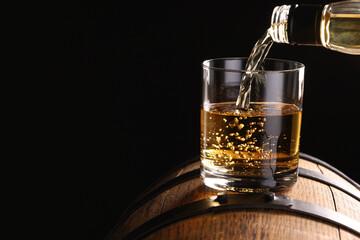 Pouring whiskey from bottle into glass on wooden barrel against dark background, closeup. Space for...