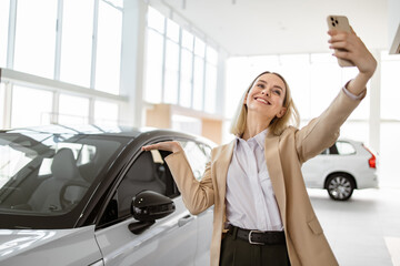 Successful businesswoman taking selfie using smartphone in car service. Happy smiling Caucasian girl buyer talking online about buying a new car.