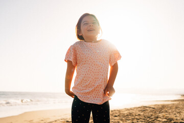 A young girl in casual wear beams with joy, basking in the warm glow of the sunset on a sandy beach