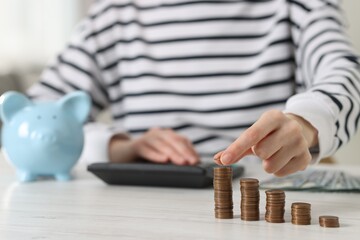 Financial savings. Woman stacking coins while using calculator at white wooden table, closeup