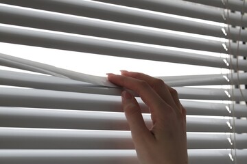 Woman separating slats of white blinds indoors, closeup