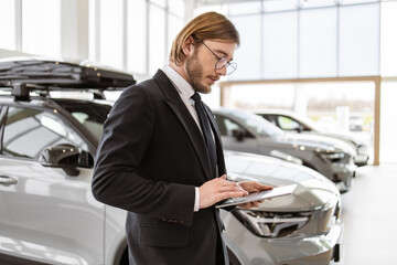 Side view of young car salesman in glasses and suit holding digital tablet. A confident bearded man...