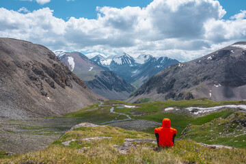 Man in vivid red jacket admire alpine scenery on sunlit green grassy hill near precipice edge. Guy on mountain pass enjoying view to few big snowy pointy peaks. Three large snow peaked tops far away.