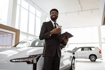 Car dealer holding clipboard in automobile showroom. Smiling salesman using smartphone while having...