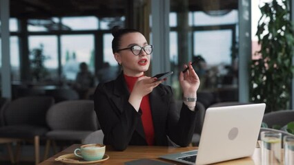 beautiful young woman in glasses and braces, a freelancer, sits in a stylish restaurant dressed in professional attire, recording a voice message on her smartphone while working on her laptop