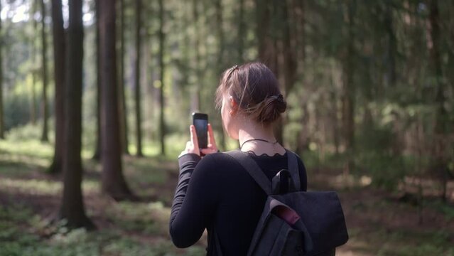 Young girl with a backpack captures the beauty of the forest on her phone. Sharing beautiful places and remembering experiences using photos in spring nature