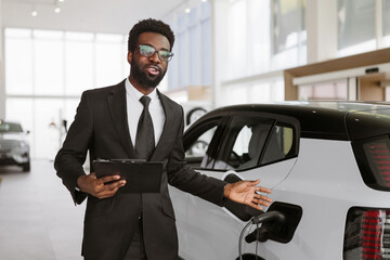 Young bearded African salesman, selling electric cars in light modern showroom with panoramic...