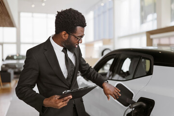 Concept of buying eco-friendly car for progressive customer. Young bearded African salesman, selling electric cars in light modern showroom with panoramic windows.