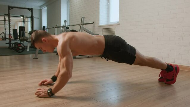 Young man in black shorts and in red sneakers in the gym performs exercises with his own weight. Discipline and self-control during systematic training in formation of beautiful muscular body