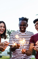 Vertical photo of happy multiracial people enjoying summer party at the beach. Diverse group of young adult students celebrating and laughing together with sparklers outdoors