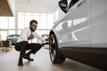 Smiling businessman wants to buy new automobile, while touch car tire wheel disk in showroom...