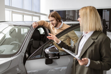 Fototapeta na wymiar Beautiful woman showing to potential buyer automobile, they looking on car cabin with opened doors. Female car dealer standing with client of dealership in showroom. Manager working with client.