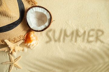 Straw hat, coconut, starfish, shells, shadow of a palm tree and the inscription SUMMER on the sand.