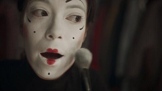 Female mime artist applying setting powder to face covered with white paint while finishing makeup for stage performance